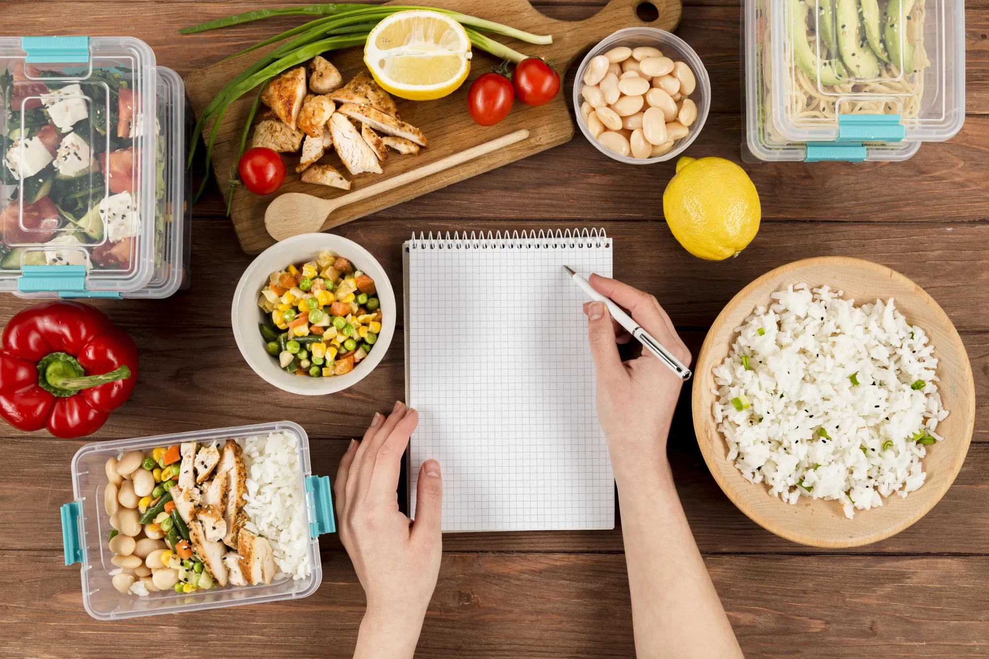A person is writing in a book with a white pen at a wooden table. Nearby are a lunchbox, a bowl, and a board with sliced chicken, lemon, and cherry tomatoes.