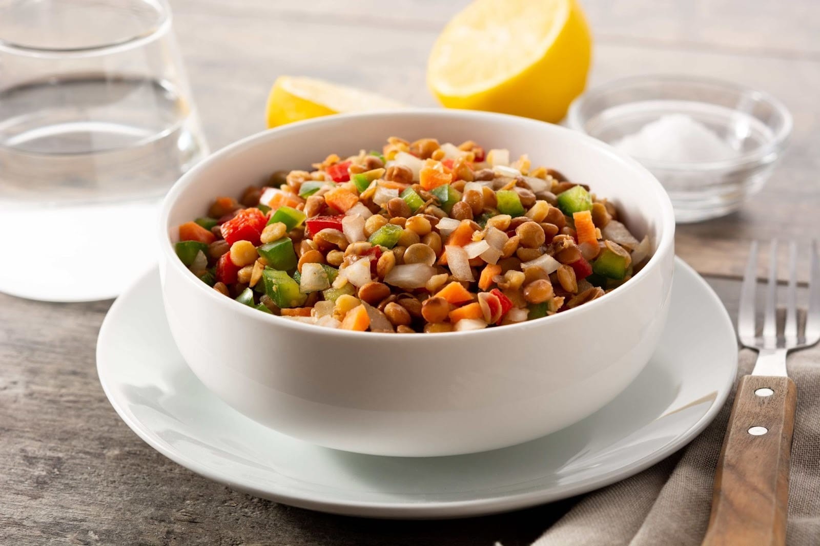 A bowl containing lentils, chickpeas, cucumbers, tomatoes and a fork, glass of water, and two lemon halves.